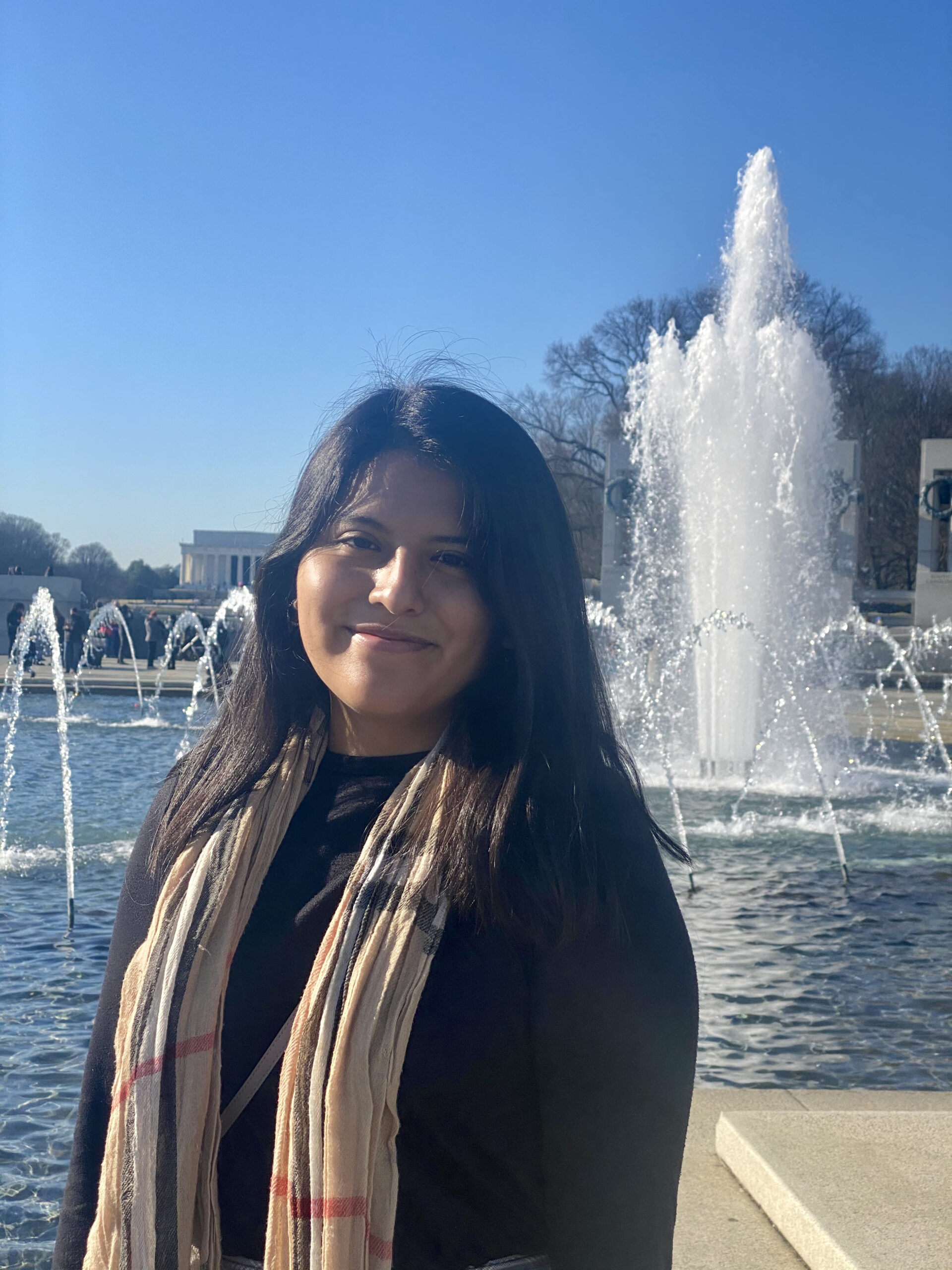 Cristina, a tan-skinned person with black hair, is outside in front of the fountains at the World War II Memorial in Washington, DC. She wears a neutral-patched scarf and smiles at the camera.