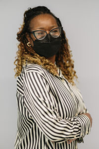 Christol, a Black woman with brownish-blonde long curly braids, and fashionable black cat-eye glasses, crosses her arms and smiles at the camera underneath her black N-95 mask. She is wearing a black and white striped button-up shirt and small gold hoop earrings. 