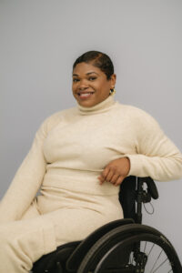 Kadesha, a medium Black woman with a short-textured pixie haircut is sitting in a black manual wheelchair leaning back on her arm, which is propped up on the push handle. She has a big smile and is wearing a cream-colored knitted turtleneck long sleeve and long pants.