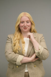 Esmerelda, a blonde light-skinned woman in her 20s, with long, wavy, platinum blonde hair is smiling at the camera with one arm crossed over her body, and one hand resting under her chin. She is wearing a white top and light brown blazer with her sleeves rolled stylishly up, in front of a grey background.