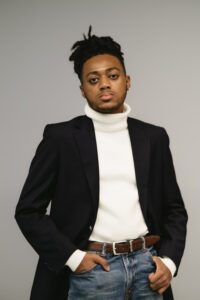 Timothy Jackson, a young Black man with locs tied up on his head, is looking with a serious expression at the camera and posing with both hands in his pockets. He is wearing a navy blazer, jeans, and a white turtleneck.