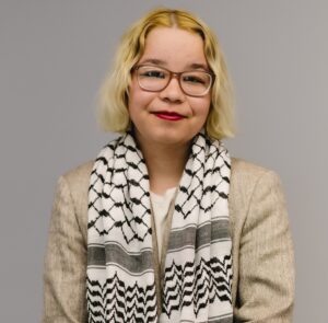 Marcy Waring, a light skinned, mixed Latina and Asian woman is looking straight at the camera. She has shoulder length blonde hair, wears brown glasses, and has red lipstick on. She is wearing a beige blazer over a white shirt. A white and black keffiyeh is over her shoulders. 