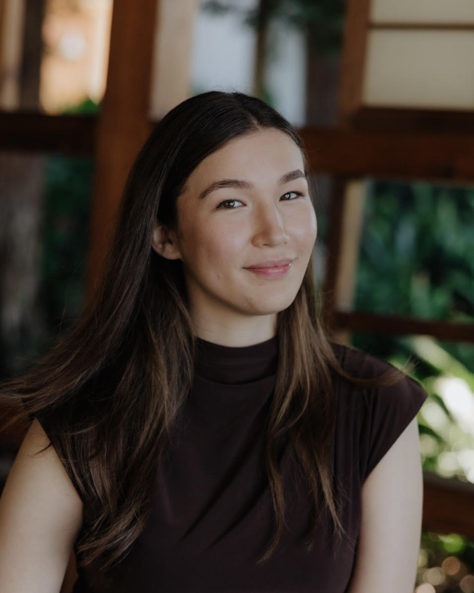 Headshot of a mixed race (White and Japanese) young woman, wearing a brown top, smiling against a green and brown background.
