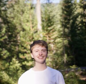 Elian, a white man with short, light-brown hair, is wearing a white t-shirt and is smiling outside on a bright, sunny morning. Behind him are a number of towering Douglas fir trees. 