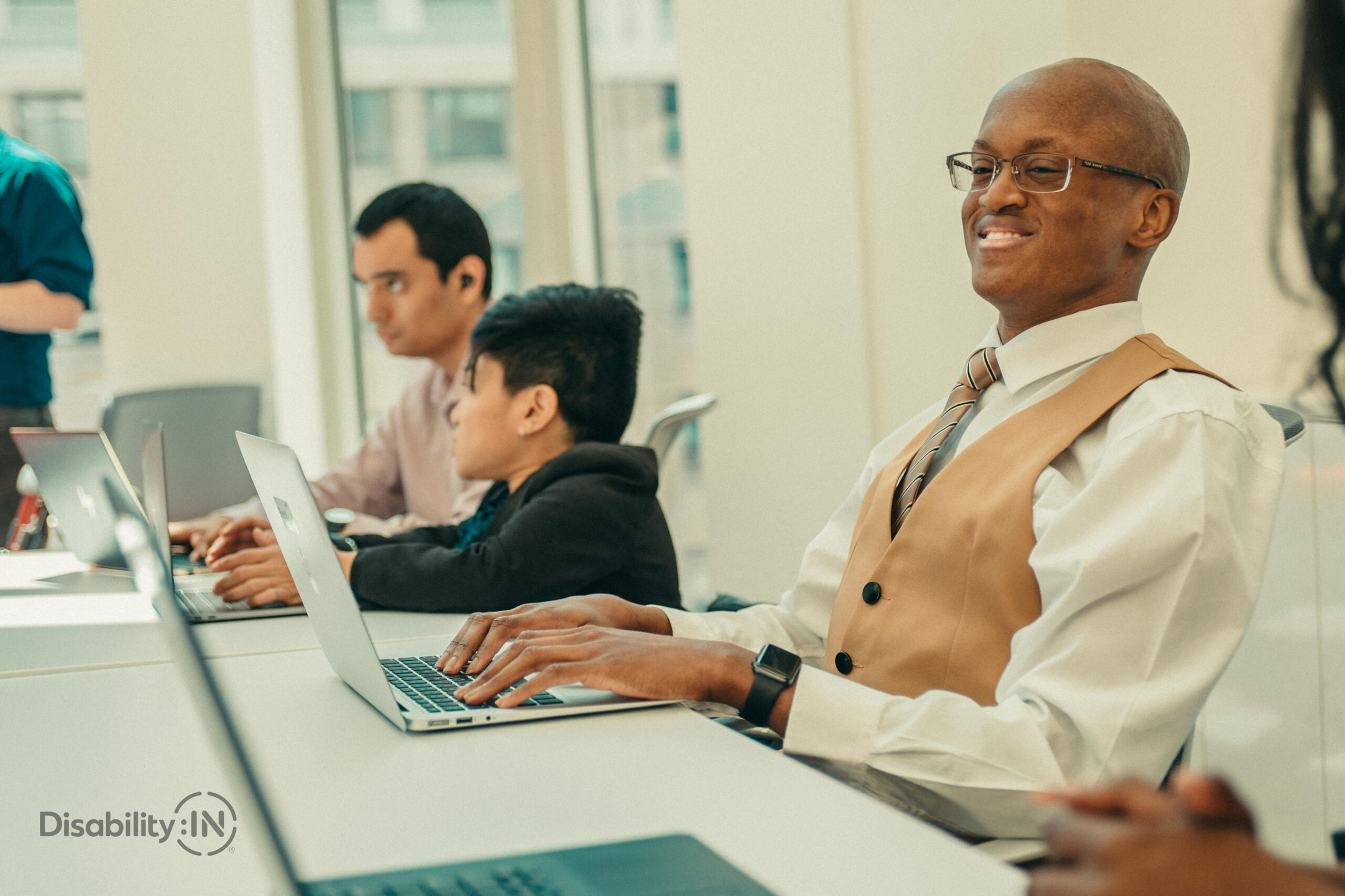Man smiles while typing on computer. Next to him are two others working and one is a wheelchair user. Credit Disability:IN