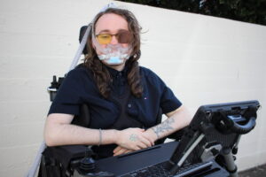ID: A white person in a power wheelchair sits in front of a white wall. The person has long wavy brown hair and is wearing glasses with an eyepatch, a BIPAP mask with a filter in front, and a dark blue button up shirt. Two abstract plant tattoos are visible on their forearm and hand resting on their belly, as well as an AAC device and keyboard.