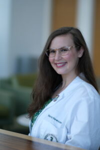 Nora is a woman with light complexion, long brown hair, and gold rimmed glasses. She is wearing a white coat.