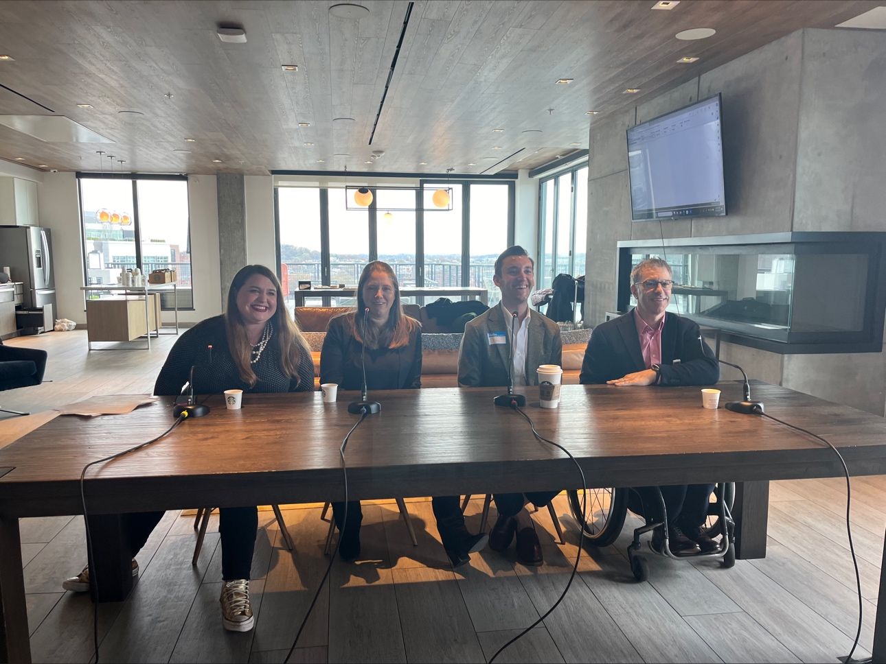 A group of four people sitting at a long wooden table with microphones and Starbucks cups in front of them. They are smiling and looking towards the camera. The setting is a modern, well-lit room with large windows and a screen on the wall in the background. One person is using a wheelchair, and the room has a professional yet casual atmosphere.