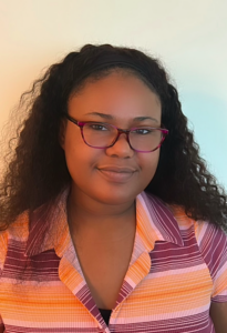 A headshot photograph of Shannon, positioned against a white background. They are shown from the shoulders up with curly hair, wearing a pink, purple, and orange button-down shirt with purple glasses.